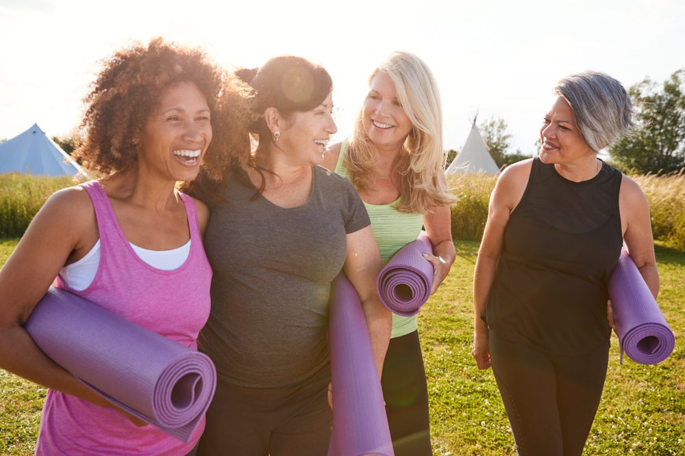 A group of women smiling together outdoors, holding yoga mats.